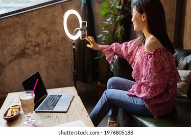 Young Woman Making Photo Or Video Content For Social Media With Smartphone And Light Of Ring Lamp. Beauty Blogger Smiles To Mobile Phone Screen. Influencer Makes Broadcast Communicates With Followers