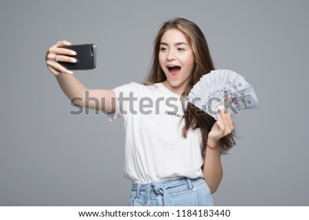 Young woman making selfie on phone while holding lots of money dollar bills isolated over gray wall