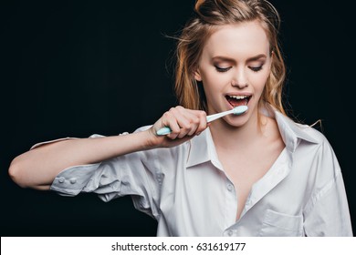 Young Woman In Male Shirt Brushing Her Teeth On Black Background, Studio Shot