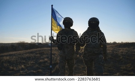 Young woman and man in military uniform going with blue-yellow banner on meadow at sunset. Female and male soldiers of ukrainian army walking with Ukraine flag at field. Resistance to russian invasion