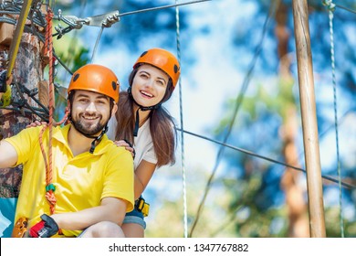 Young woman and man in protective gear are sitting on rope bridge hanging on high trees, posing and smiling. Rope adventure park with obstacles and ziplines. Extreme rest and summer activities concept - Powered by Shutterstock