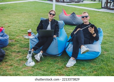 A young woman and a man are sitting in a park on a bean bag and smoking hookah. Happy couple relaxing outdoors with hookah - Powered by Shutterstock
