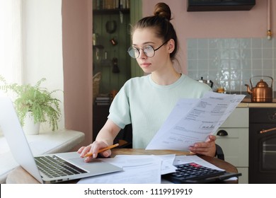 Young woman managing domestic budget, sitting at kitchen table with open laptop, documents and calculator, using touchpad, making notes with pencil