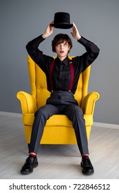 Young woman in man's suit sitting in yellow armchair and holding a hat above her head