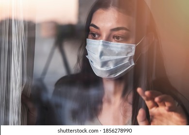 Young Woman With Mask Looking Out The Window During Home Confinement. With The City Reflected In The Glass. Looking Forward To Going Outside. Concept Covid 19.