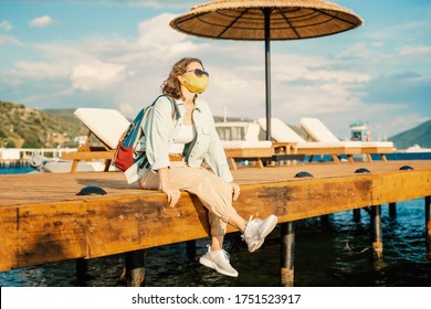 Young Woman With A Mask On Her Face Sits On A Pier By The Sea, Summer Travel Trend 2020, Health Safe Vacation