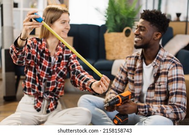 Young Woman With Measure Tape Looking At Smiling Man With Electric Drill While Sitting Together On Floor. Multiracial Couple Measuring Free Space For New Furniture At Modern Apartment.