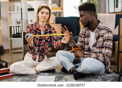 Young Woman With Measure Tape Looking At Smiling Man With Electric Drill While Sitting Together On Floor. Multiracial Couple Measuring Free Space For New Furniture At Modern Apartment.