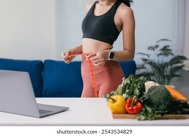 Young woman measuring her waist with a tape measure while standing near a table with fresh vegetables and a laptop, promoting a healthy lifestyle at home - Powered by Shutterstock