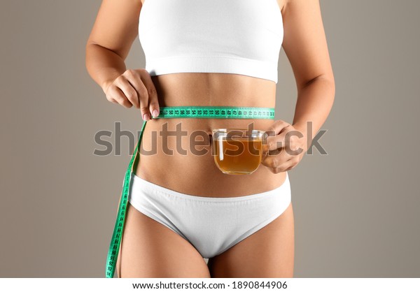 Young woman with measuring tape\
holding cup of diet tea on beige background,\
closeup