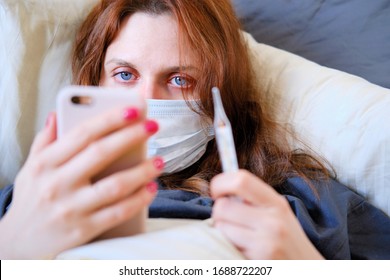A young woman in a medical mask calls on the phone to call a doctor at home. Redhead girl sick with coronavirus lies in a home bed with a thermometer and a phone in her hands - Shutterstock ID 1688722207
