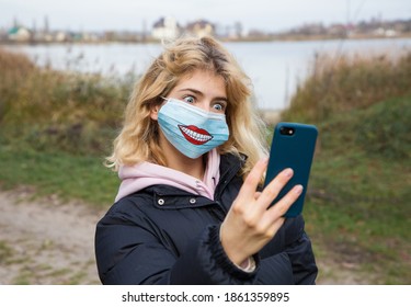 young woman in medical mask with drawn cheerful smile makes  selfie portrait, funny rounded eyes. holds  phone in front of her. Social isolation, walks in  fresh air during  covid-19 pandemic - Shutterstock ID 1861359895