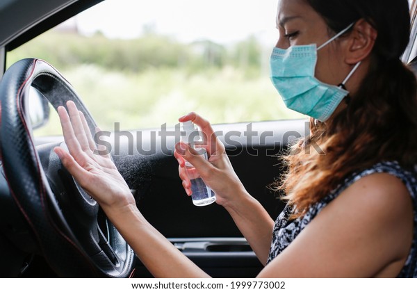 Young woman in a medical mask driving a car\
cleaning hands with alcohol spray, coronavirus protective mask,\
driver on city road during coronavirus outbreak. Safety during a\
covid-19 epidemic.