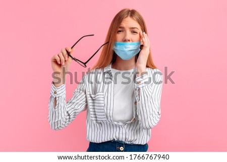 young woman with a medical mask on her face and glasses with poor vision tries to read a text on a pink background