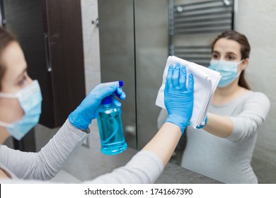 Young Woman With Medical Mask Polishing Mirror With Rag And Using Disinfectant Spray. Housekeeping And Cleaning Service.