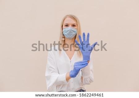 Young woman in a medical mask puts on protective blue latex gloves on her arm, isolated on beige background, protection against coronavirus