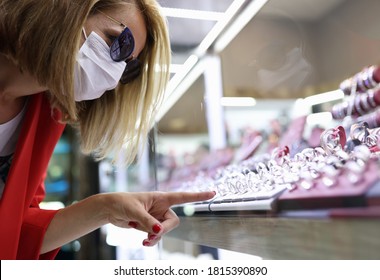 Young Woman In Medical Mask, Sunglasses, Red Jacket Points Her Finger At Jewelry Display. Social Distance And Covid 19 Etiquette Concept.