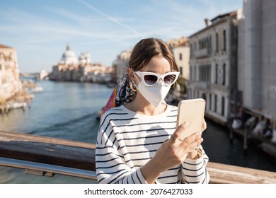 Young Woman In Medical Mask Traveling During Pandemic In Italy. Concept Of Social Rules Of Wearing A Mask During A Pandemic. Woman Using Smartphone While Standing On The Bridge In Venice