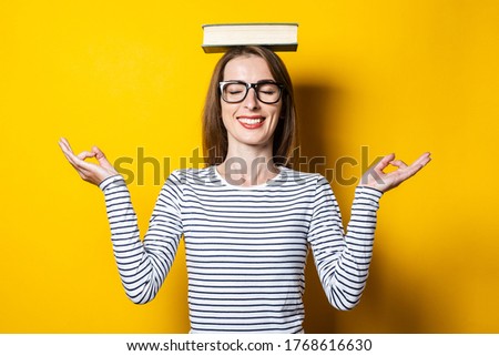 Young woman meditates with closed eyes holds a book on her head on a yellow background.