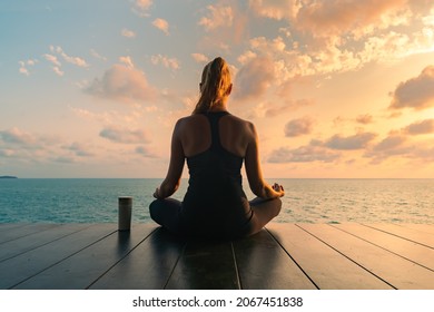 Young woman meditating in a black clothes for yoga  on a wooden deck on the top by the sea. There is a thermo mug nearby.On the background  colorful sunrise sky and sea view.