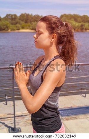 Young woman meditating in city by the river in summer, closed eyes, hands together.
