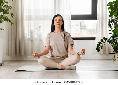 Young woman meditating sitting on a yoga mat near a big window - Shutterstock ID 2304884285