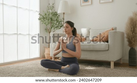 a young woman meditating in a yoga pose, with closed eyes and hands in prayer position. The minimalist home environment with soft natural light complements her peaceful expression