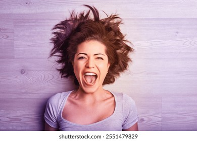 Young woman with messy brown hair lies on a wooden floor, her head tilted back and mouth wide open in a scream of frustration, joy, or excitement - Powered by Shutterstock