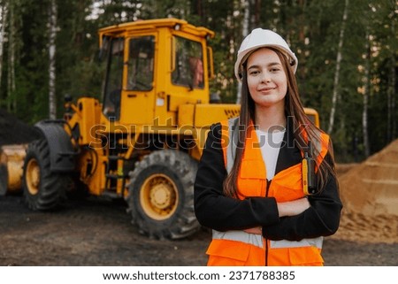 A young woman mine worker is standing in front of a large dump truck or grader. Looking at camera.