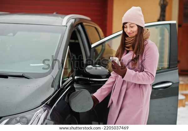 Young woman with mobile phone at hand charging\
her electric car in\
winter.