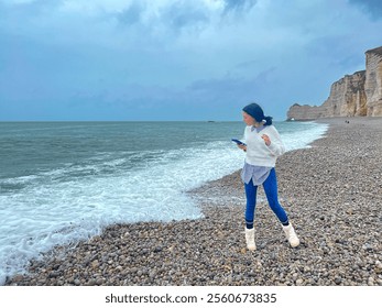 Young woman with mobile phone in her hands walking on the beach of the sea at Etretat, Normandy, France.  - Powered by Shutterstock