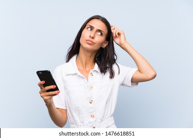 Young woman with a mobile phone over isolated blue wall having doubts and with confuse face expression