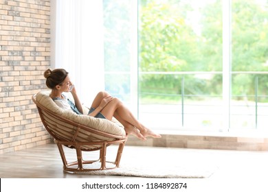 Young woman with mobile phone sitting in papasan chair near window at home