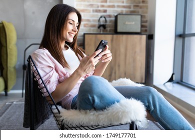 Young Woman With Mobile Phone Sitting In Papasan Chair Near Window At Home