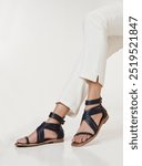 Young woman modeling stylish Mediterranean-inspired leather gladiator sandals in dark blue, paired with white cropped trousers. The scene evokes quiet luxury, emphasizing minimalist, sophisticated.