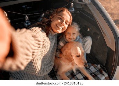 A young woman mother with her cute daughter and retriever dog sitting on a warm blanket in the open trunk of a car and taking a selfie on the phone. Family traveling with a pet. - Powered by Shutterstock