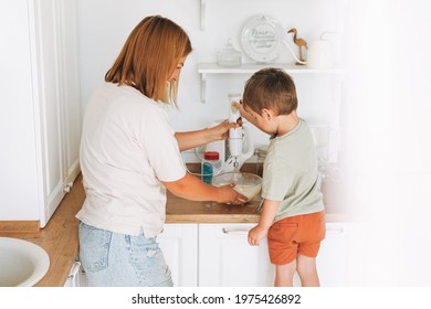 Young Woman Mother And Her Toddler Boy Son Cooking With Blender At The Table In Kitchen At Home