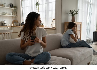 Young Woman Mother Holding Smartphone Looking At Offended Upset Teen Girl Daughter Sitting Separately On Sofa. Sad Girl Child Expecting Attention And Time From Parent Spending Too Much Time On Phone