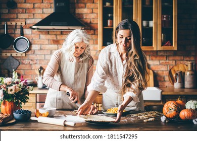 Junge Frau und Mutter in weißen Schränken stehen am großen braunen Küchentisch und bereiten ein leckeres Abendessen zu. Senior Mutter und glückliche erwachsene Tochter backen Kürbiskuchen zusammen zu Hause.