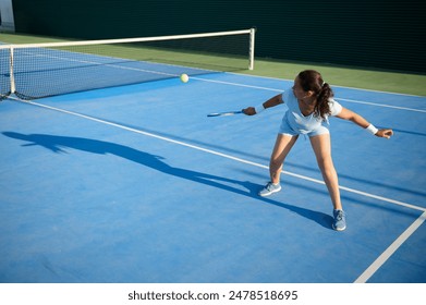 A young woman in motion hitting a tennis ball during a game on an outdoor blue tennis court, displaying agility and determination. - Powered by Shutterstock