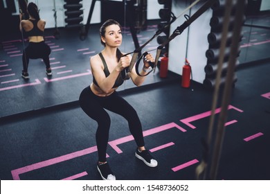 Young Woman Motivated For Getting Stronger Training In Gym With TRX Equipment And Doing Squat Exercises For Butt And Legs 