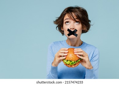 Young Woman With Mouth Sealed With Tape Wearing Sweater Holding Burger Want To Eat Look Overhead Isolated On Plain Pastel Light Blue Background Studio Portrait. People Lifestyle Diet Junk Food Concept