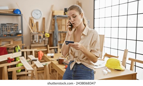 A young woman multitasking on the phone while holding a credit card in a carpentry workshop. - Powered by Shutterstock