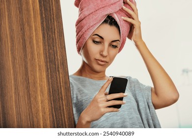 Young woman multitasks at home, holding her smartphone, reading news, and drying her hair with a towel after a shower, embodying a relaxed and comfortable modern lifestyle - Powered by Shutterstock