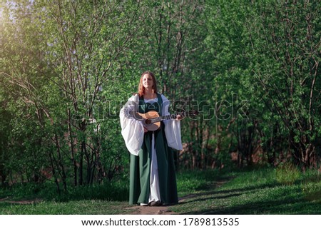 Young woman musician in medieval long dress walks with guitar in sunny summer green forest. Fantasy girl or bard at summer nature.