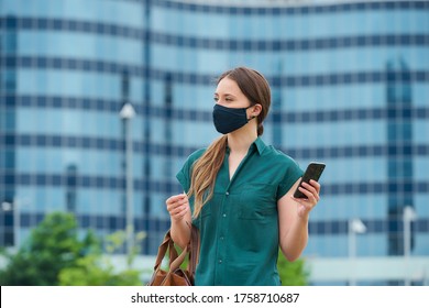 A young woman in a navy blue medical face mask to avoid the spread of coronavirus is holding her leather bag in the center of the city. A girl with long hair is using a smartphone in downtown. - Powered by Shutterstock