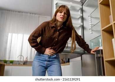 Young Woman Near Empty Refrigerator Feels Pain In Her Stomach. Hungry Girl Holding On To Her Abdomen