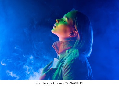 Young woman in neon multi-colors light on smoke, steaming background. Millennial enigmatic futuristic lady in fashion trench coat, stylish outfit 