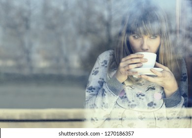 Young Woman In Night Dress Drinking Warm Coffee Or Tea  Inside Home Looking Out Through The Window Glass On Cold Winter Morning. Concept Of Lazy Winter Morning And Comfort Of A Cozy Appartment.