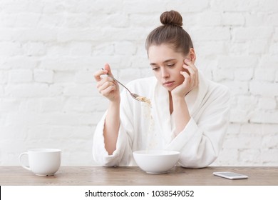 Young woman with no appetite, tired, bad mood and sleepy, sitting in the white loft kitchen, having unsavory unpalatable unappetizing breakfast - Powered by Shutterstock